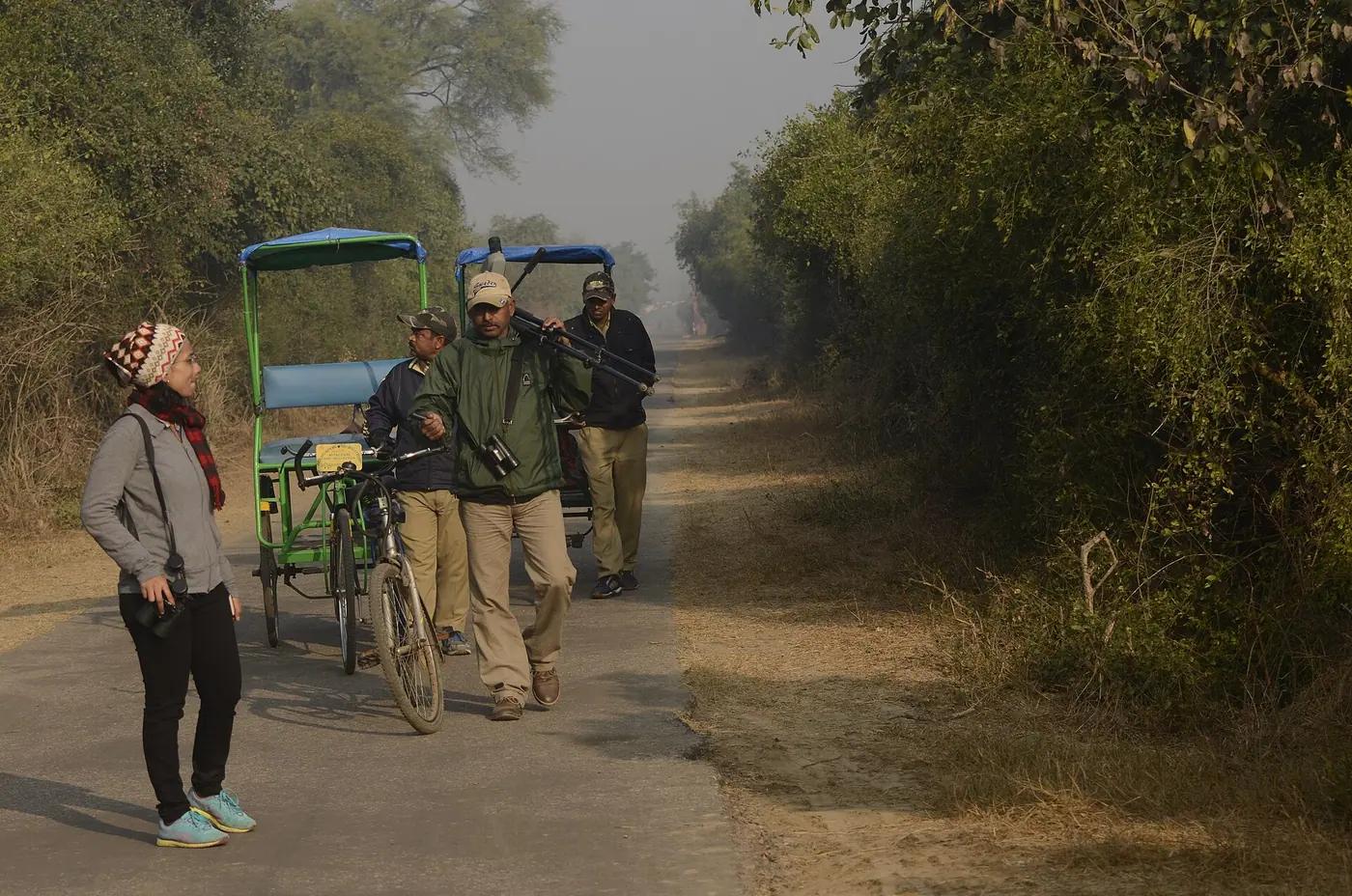 Local bird guides carrying a spotting scope and binoculars beside cycle rickshaws on a reserve track, with a visitor listening