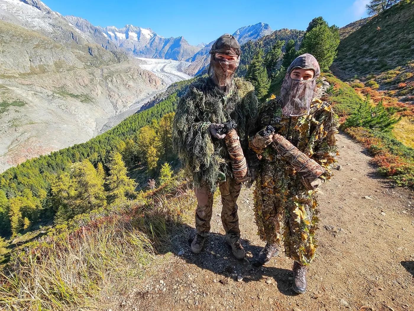 Two wildlife photographers in ghillie suits holding cameras with long lenses on a mountain trail above a glacier