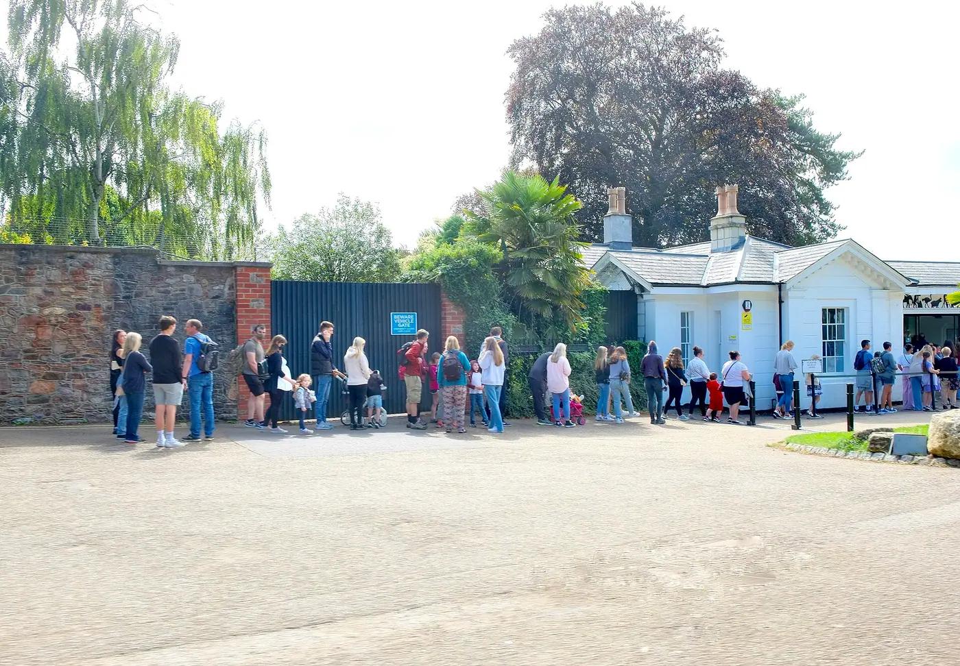 Families queuing along a stone wall at the entrance gate of a zoo on a sunny day