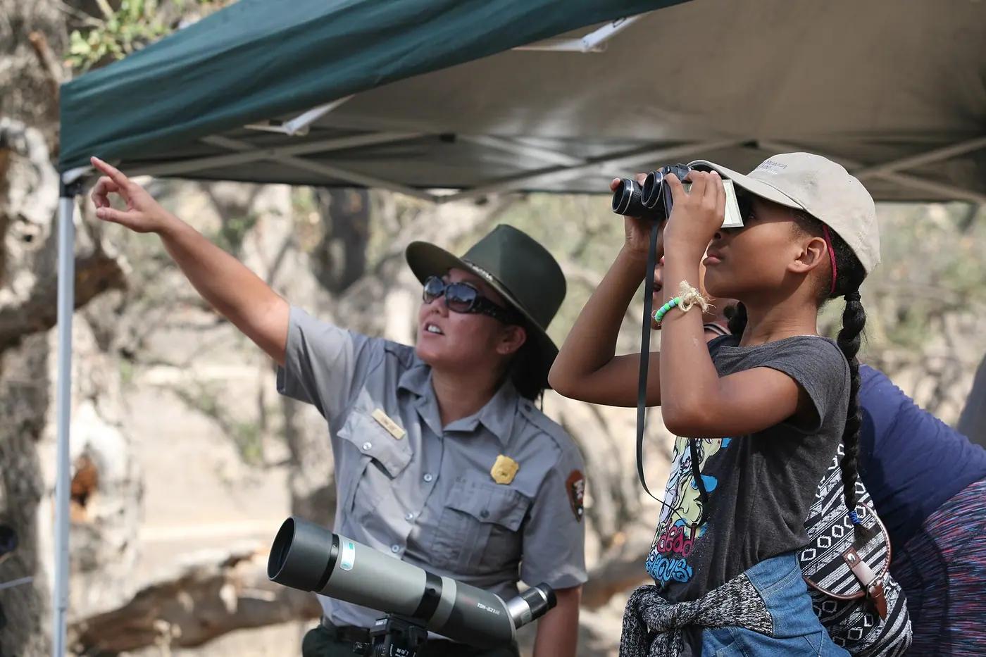 A ranger points upward while a young guest beside her looks through binoculars during a bird walk