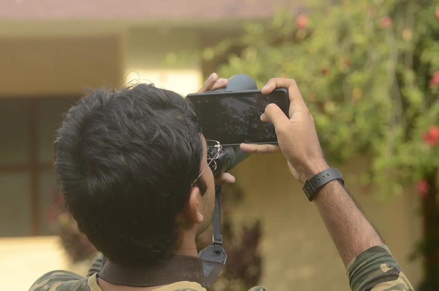 A birdwatcher seen from behind holding a phone to the eyepiece of binoculars to photograph a bird