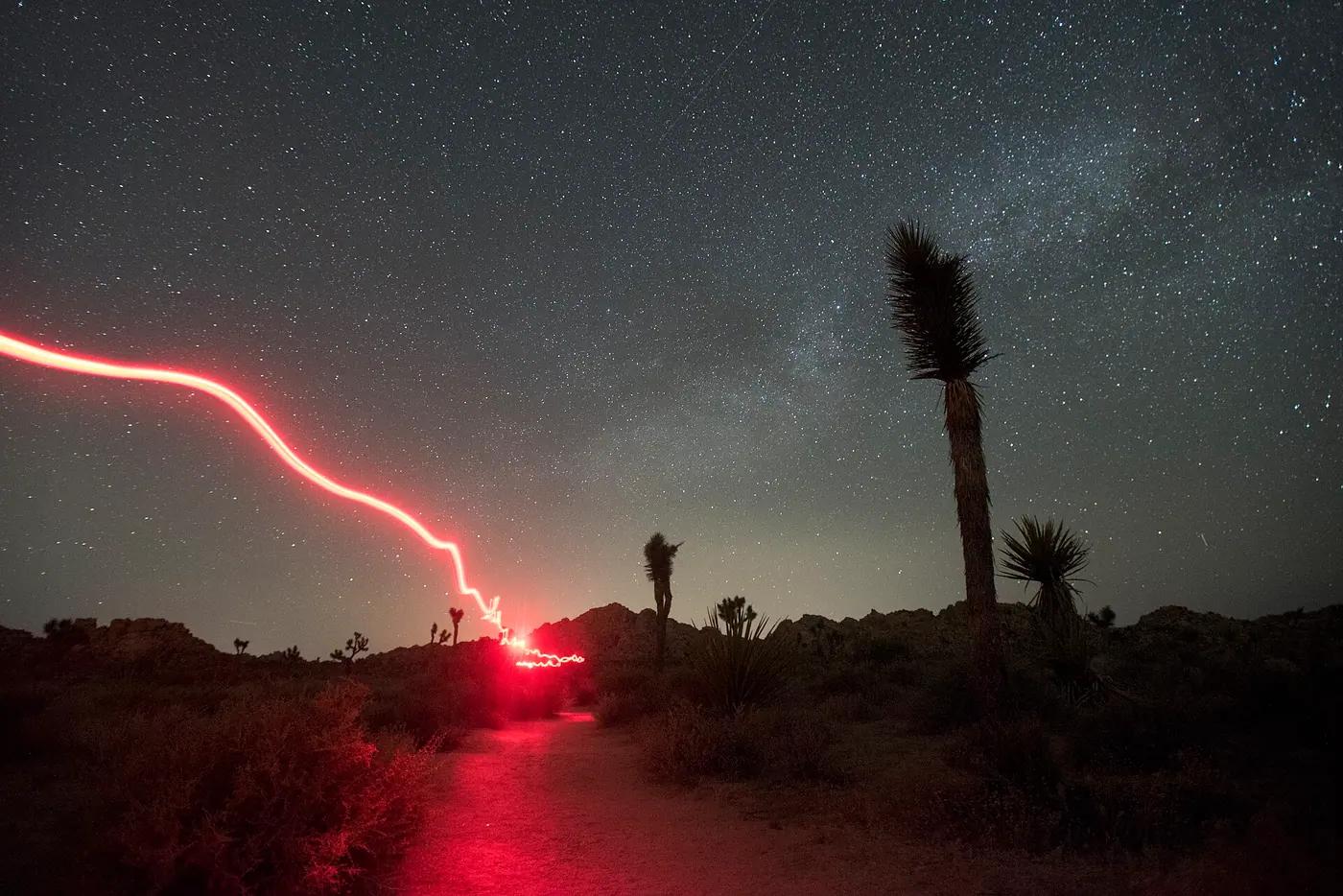 A long-exposure of a hiker's red headlamp tracing a desert trail under a starry sky