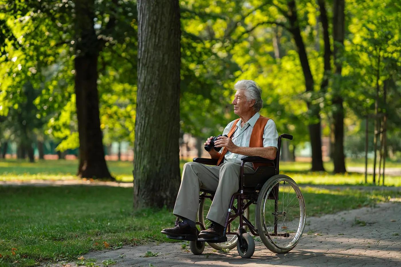 An older man in a wheelchair holding binoculars on a paved path under park trees