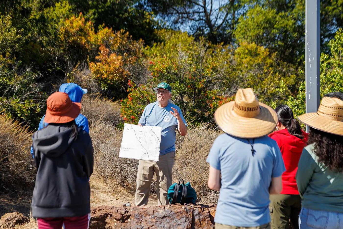 A guide holding a site map talks to a small group in sun hats on a scrubland trail