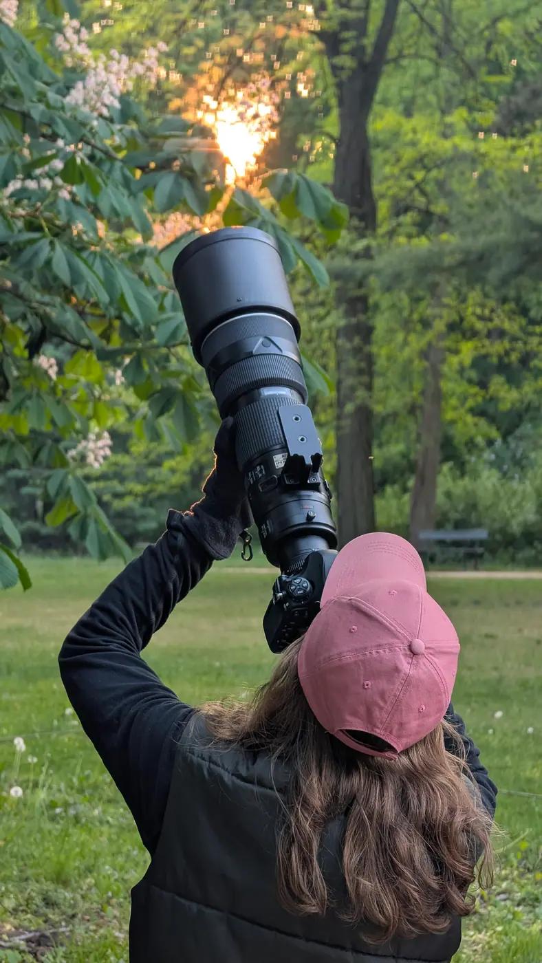 A photographer in a pink cap holds a camera with a large 200-400mm telephoto lens up toward trees at sunset