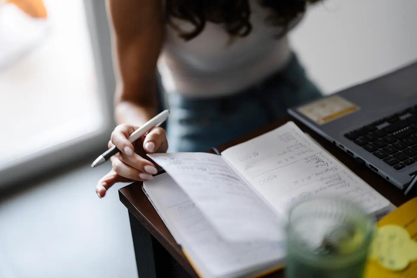 A person writing in an open notebook next to a laptop